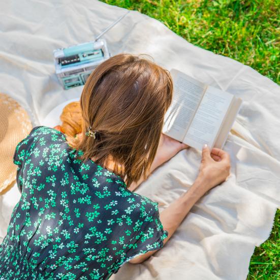 Kvinde, der lægger på et tæppe og læser en bog udenfor.