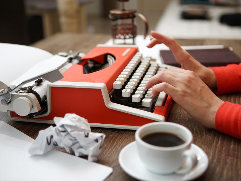 Hænder, der skriver på skrivemaskine. Rundt om er en kop kaffe og noget sammenkrøllet papir.