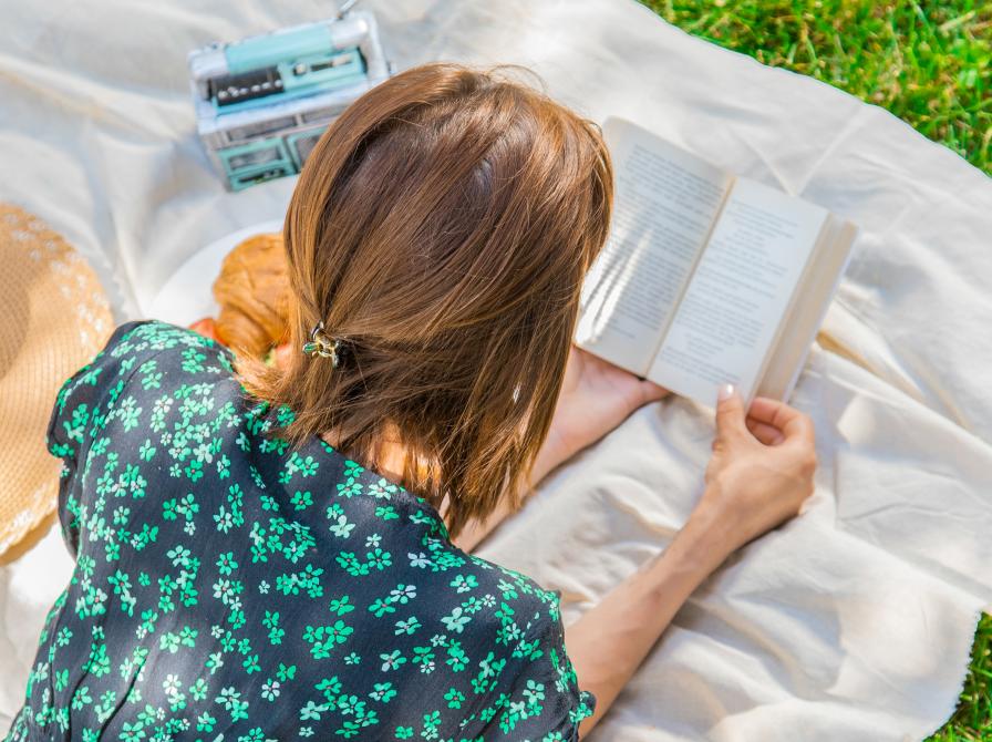 Kvinde, der lægger på et tæppe og læser en bog udenfor.